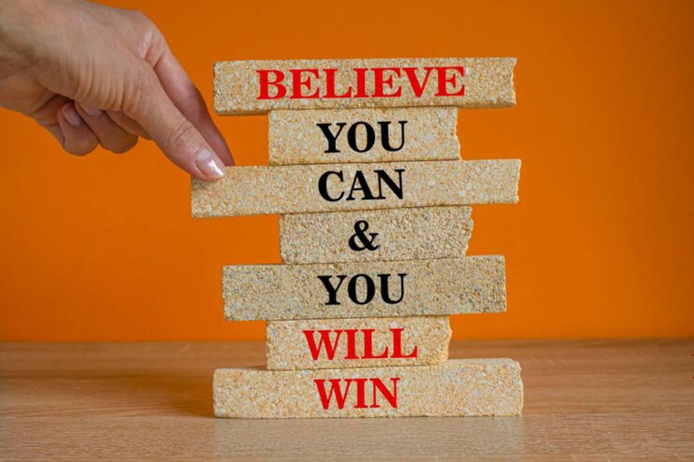 A hand supports stacked blocks with motivational phrases: 'BELIEVE,' 'YOU' 'CAN,' & 'YOU WILL WIN,' against an orange background.