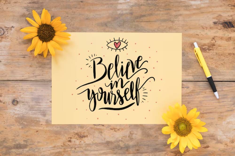 Handwritten 'Believe in yourself' on beige paper, surrounded by sunflowers and a pen, on a rustic wooden table.