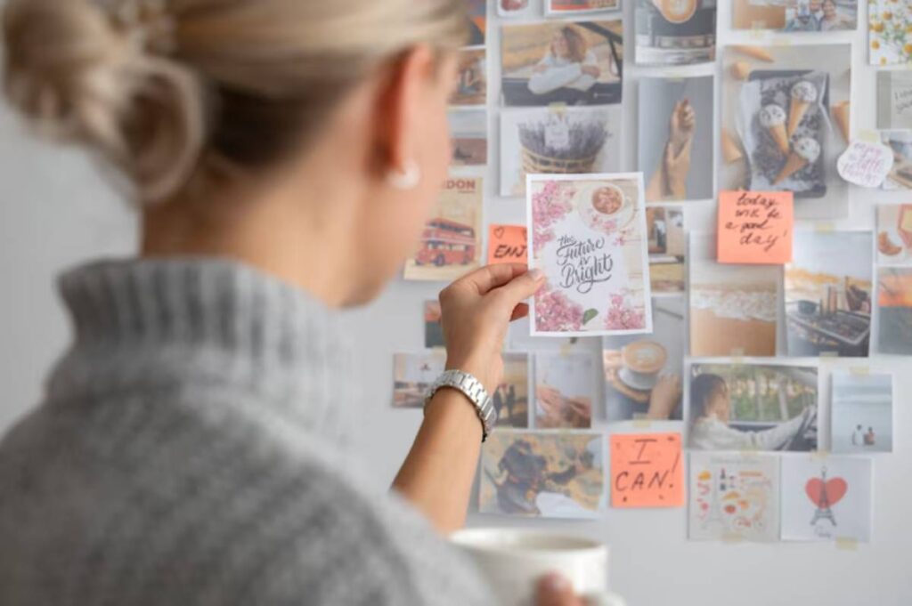 A woman in a gray sweater holds a motivational card reading 'The Future is Bright,' gazing at a vision board filled with images.