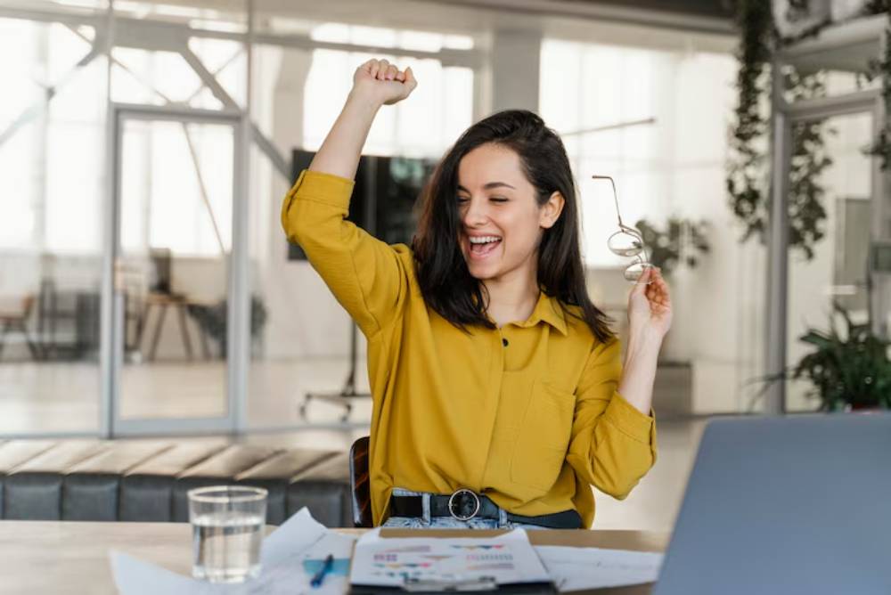 A person in a yellow shirt raises one fist triumphantly, holding glasses in the other hand, surrounded by office papers and a laptop.