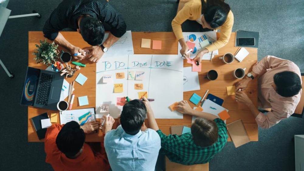 A diverse group of professionals collaborate at a table with sticky notes, charts, and laptops, organizing tasks into categories.