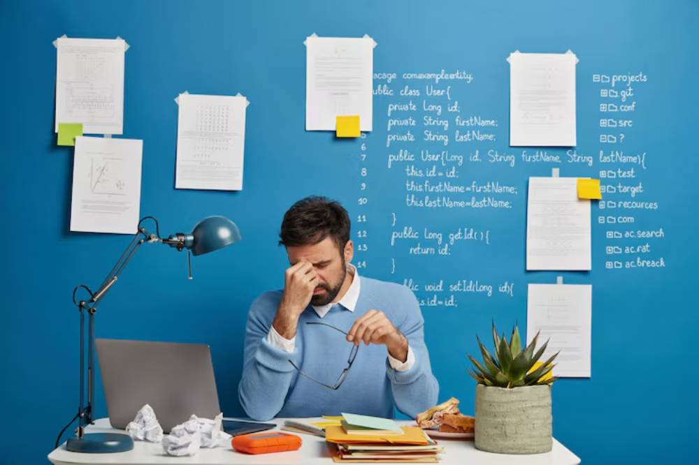 A person sits at a desk cluttered with papers and a laptop, against a blue wall filled with notes and coding fragments, with a plant nearby.