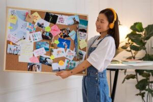 A person holds a corkboard filled with colorful photos, notes, and stickers, showcasing travel and creative inspirations in a bright room.