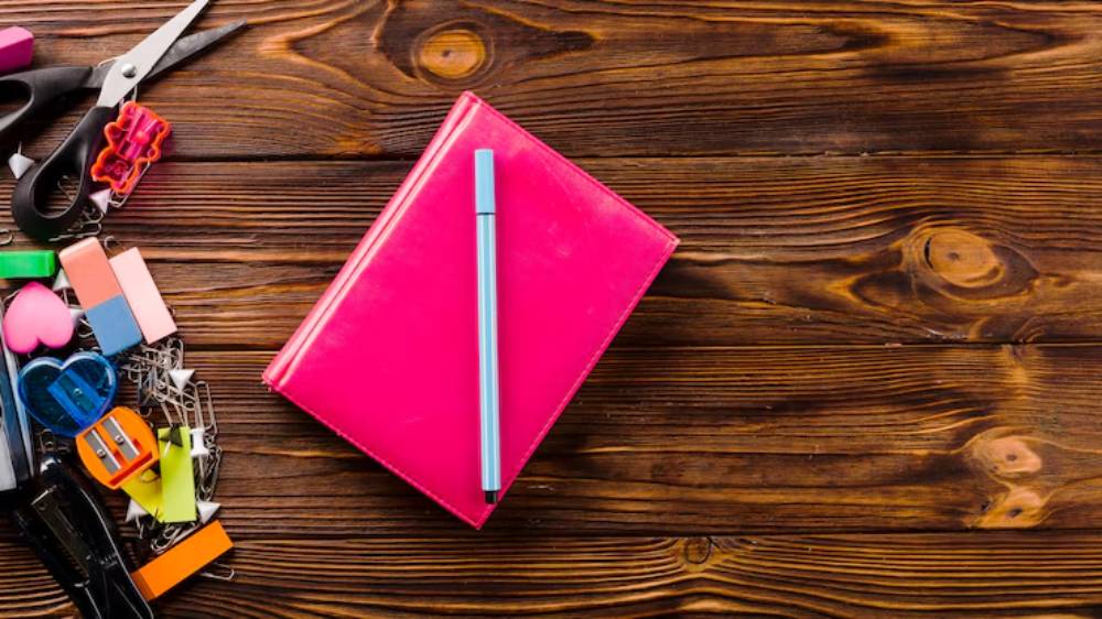 A pink notebook with a blue pen rests on a wooden table surrounded by colorful stationery supplies like scissors and paper clips.
