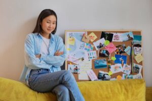 A young person sits on a yellow couch, arms crossed, next to a colorful vision board filled with photos and inspirational notes.