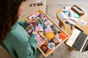 A person creates a vision board filled with colorful images and notes, surrounded by crafting supplies and a laptop on a coffee table.