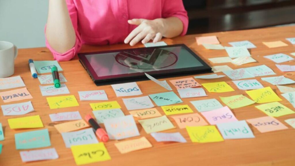 A person in a pink shirt gestures over a tablet surrounded by colorful sticky notes on a wooden table, indicating brainstorming activity.