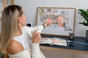 A woman in a cozy setting points to a vision board filled with images and notes, while holding a cup in her other hand.