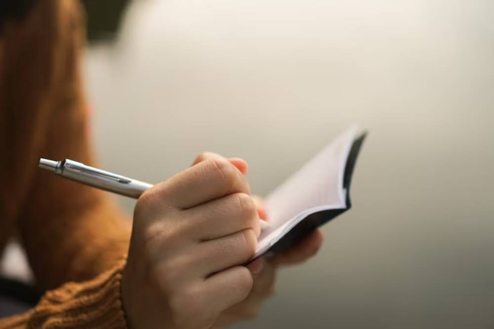 A person in a brown sweater writes in a small notebook with a silver pen, surrounded by a blurred, soft background.