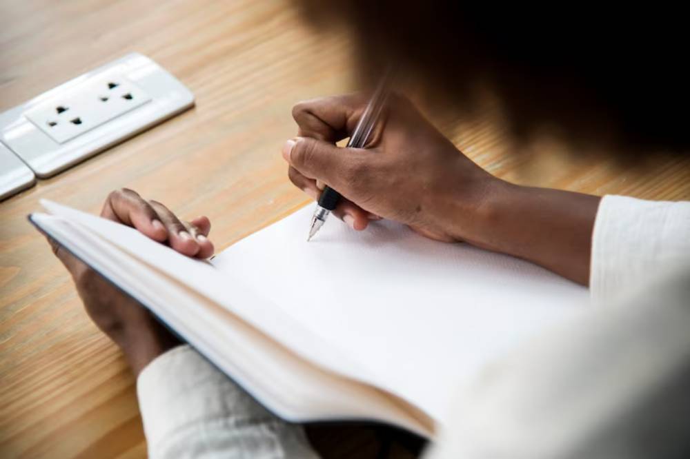 A person writes in a blank notebook with a pen, seated at a table with electrical outlets in the background.
