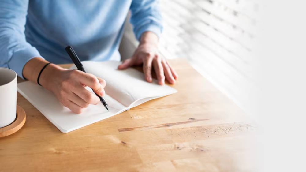 A person in a blue sweater writes in a blank notebook on a wooden table, with a cup of coffee nearby. Natural light filters through blinds.