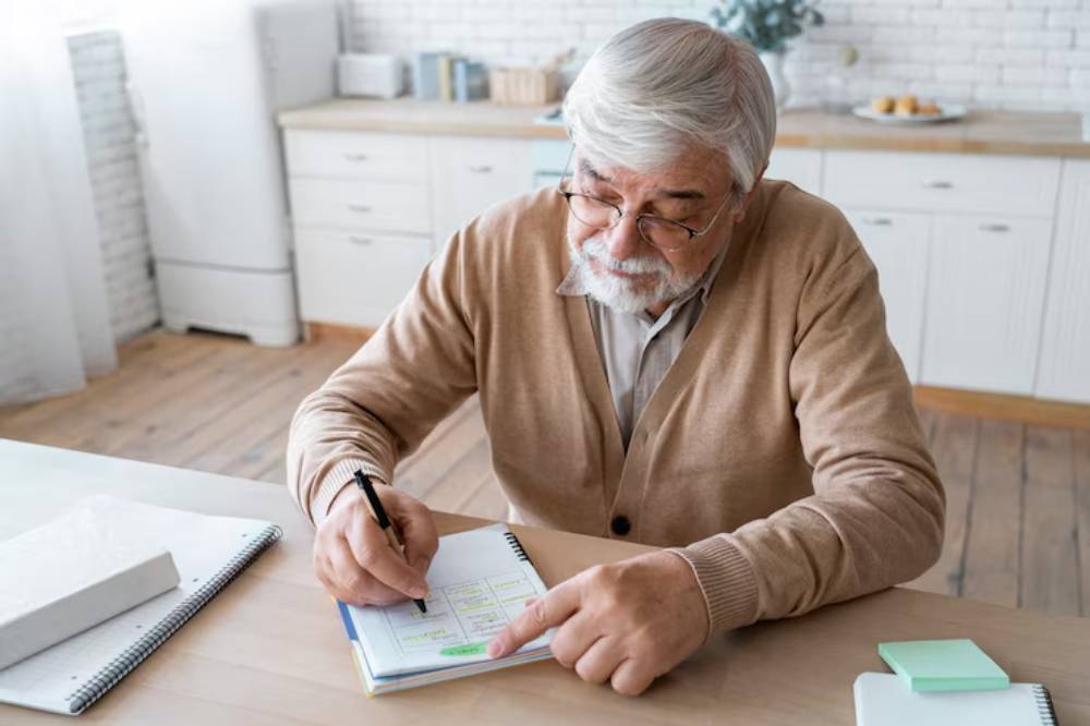An elderly person in a beige cardigan is writing in a notebook at a wooden table in a bright, modern kitchen.