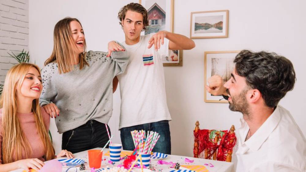 A group of four young people gather around a colorful party table decorated with vibrant drinks and festive decorations, smiling and posing.