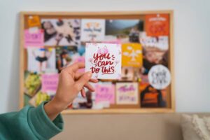 A person holds a motivational card reading 'You Can Do This' in front of a vibrant vision board filled with photos and notes.
