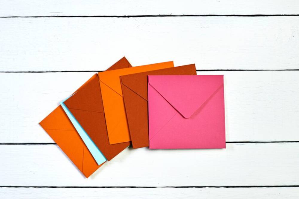 A stack of colorful envelopes in orange, brown, and pink on a white wooden surface, showcasing various shades and sizes. 