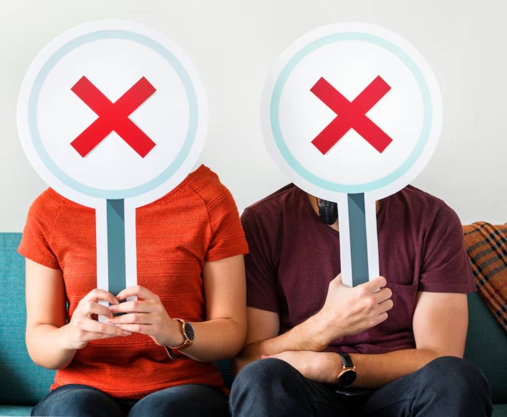 A woman in an orange shirt and a man in a maroon shirt hold large signs with red 'X' marks covering their faces, sitting on a couch.