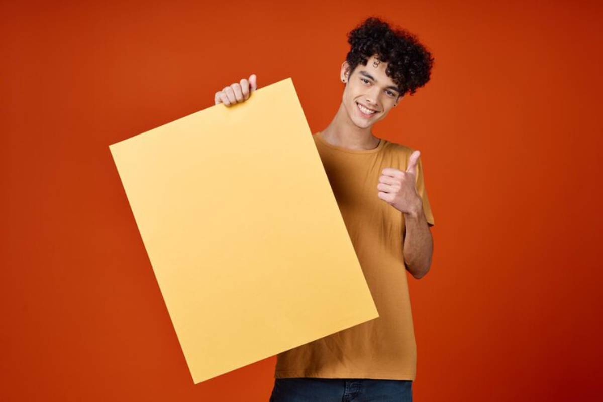 A person in a mustard-colored shirt holding a large yellow poster board and giving a thumbs up, set against an orange background.