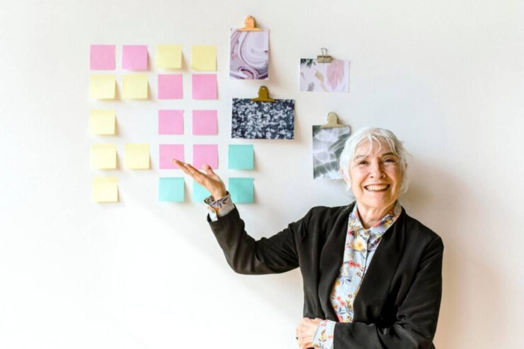 A person in a black blazer stands beside a wall covered with colorful sticky notes and artistic prints, gesturing towards the notes.