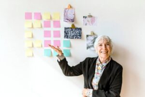 A person in a black blazer stands beside a wall covered with colorful sticky notes and artistic prints, gesturing towards the notes.