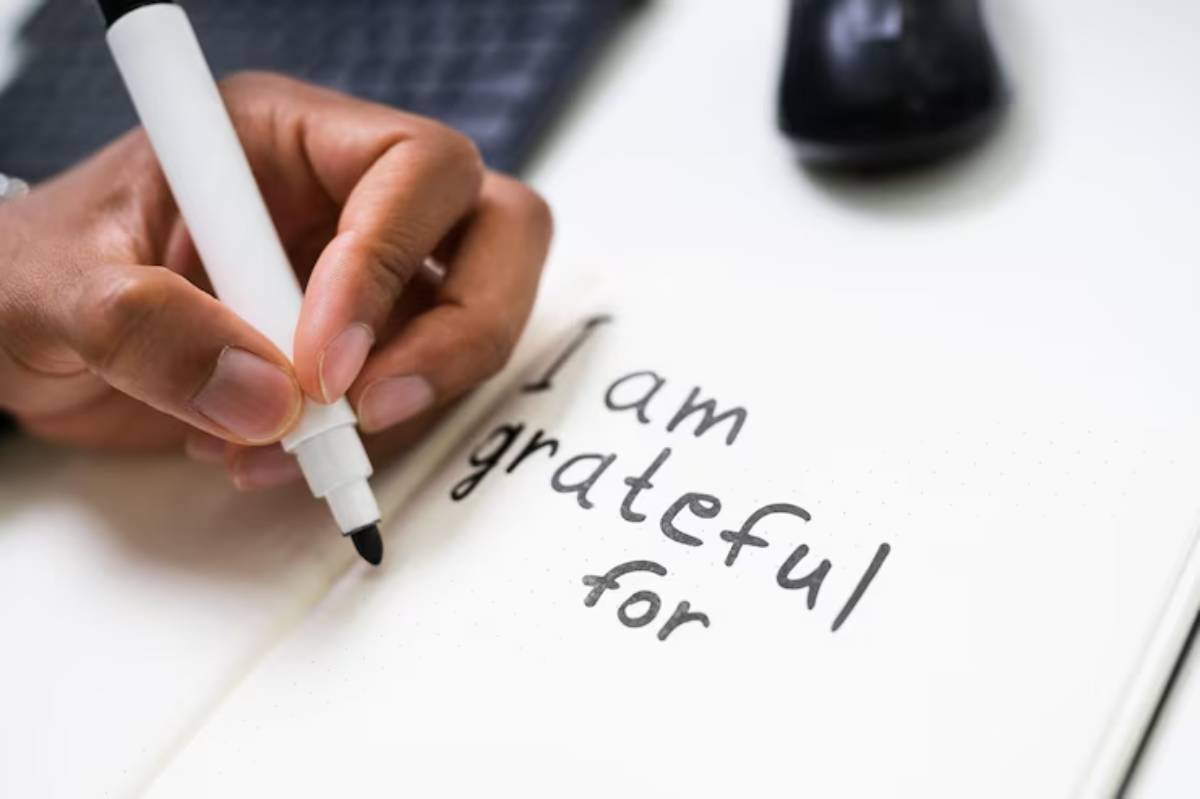 A person's hand holding a white marker writes 'I am grateful for' in a notebook, with a computer mouse and a planner in the background.