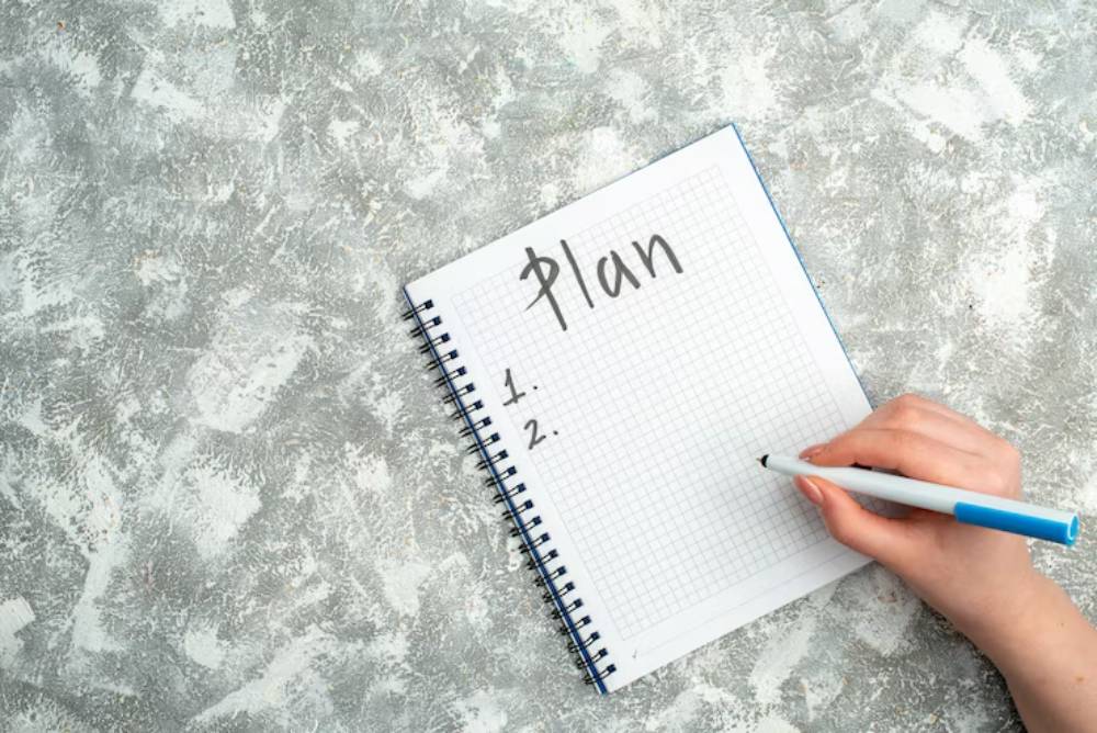 A hand holding a blue pen, writing a numbered list titled Plan in a spiral notebook on a textured grey surface.