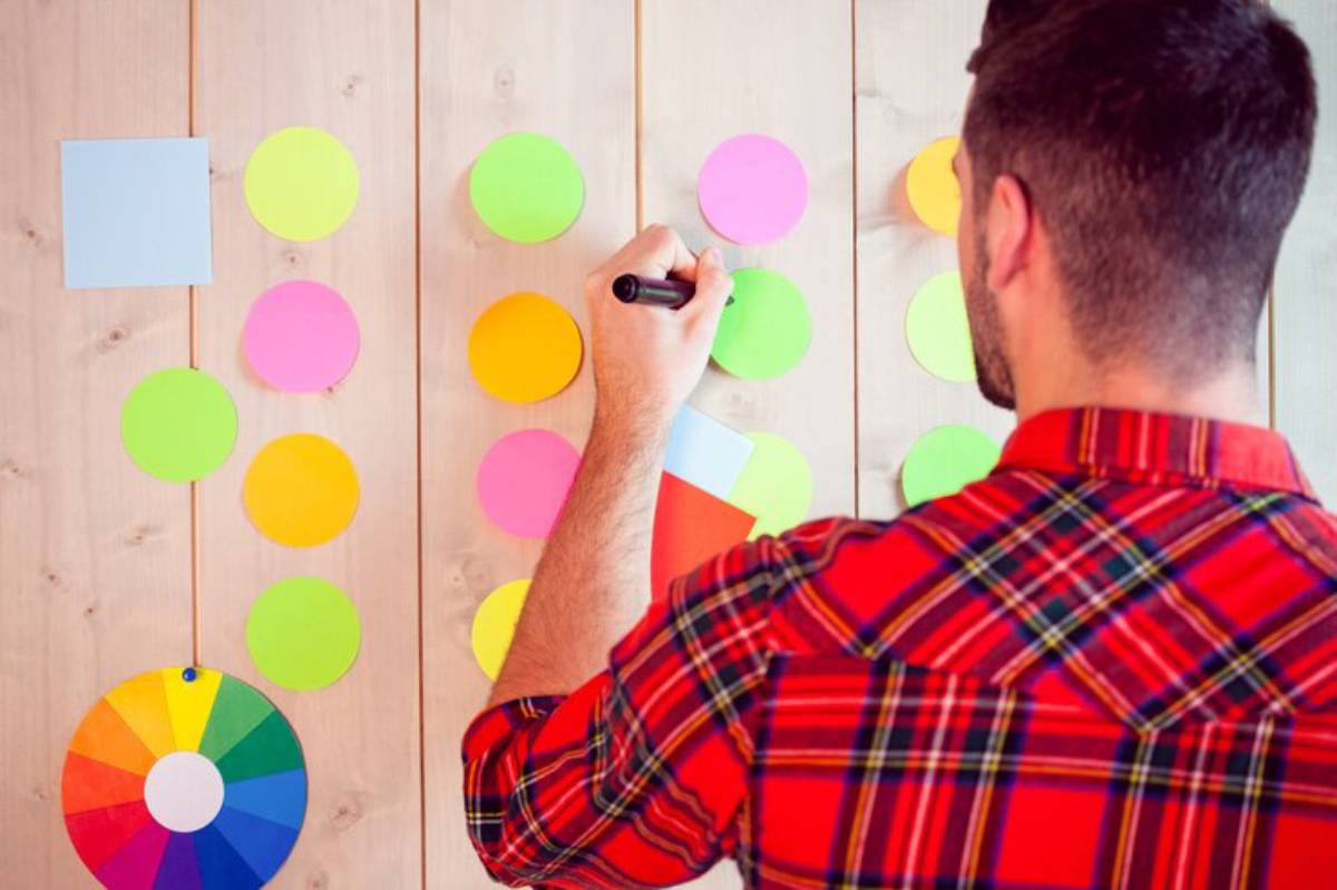 A man in a plaid shirt writes on colorful sticky notes arranged on a wooden wall, with a color wheel at the bottom left.