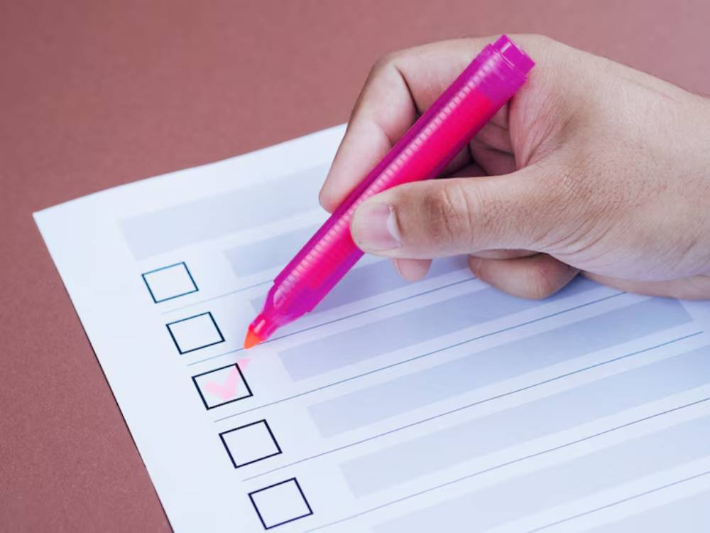 A hand holds a pink highlighter, marking a checkbox on a blank checklist paper against a brown background.