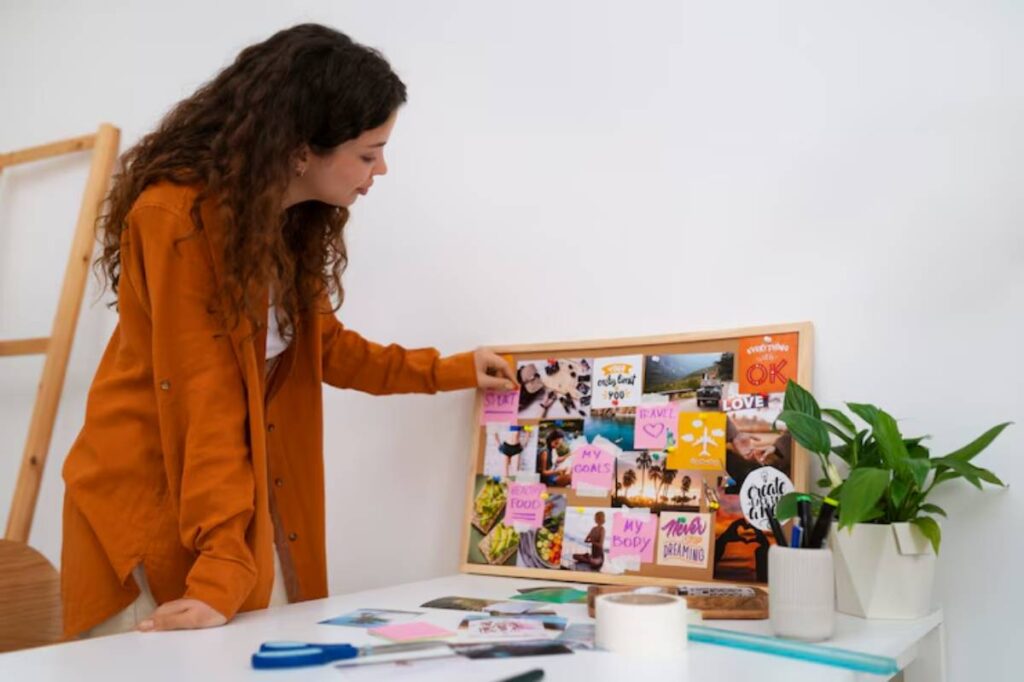 A person in an orange shirt arranges a vision board filled with motivational images and notes on a white table, surrounded by plants.