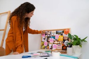 A person in an orange shirt arranges a vision board filled with motivational images and notes on a white table, surrounded by plants.