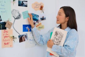 A person wearing a polka-dot shirt stands near a motivational vision board collage filled with notes, photos, and art, holding a sketchbook.