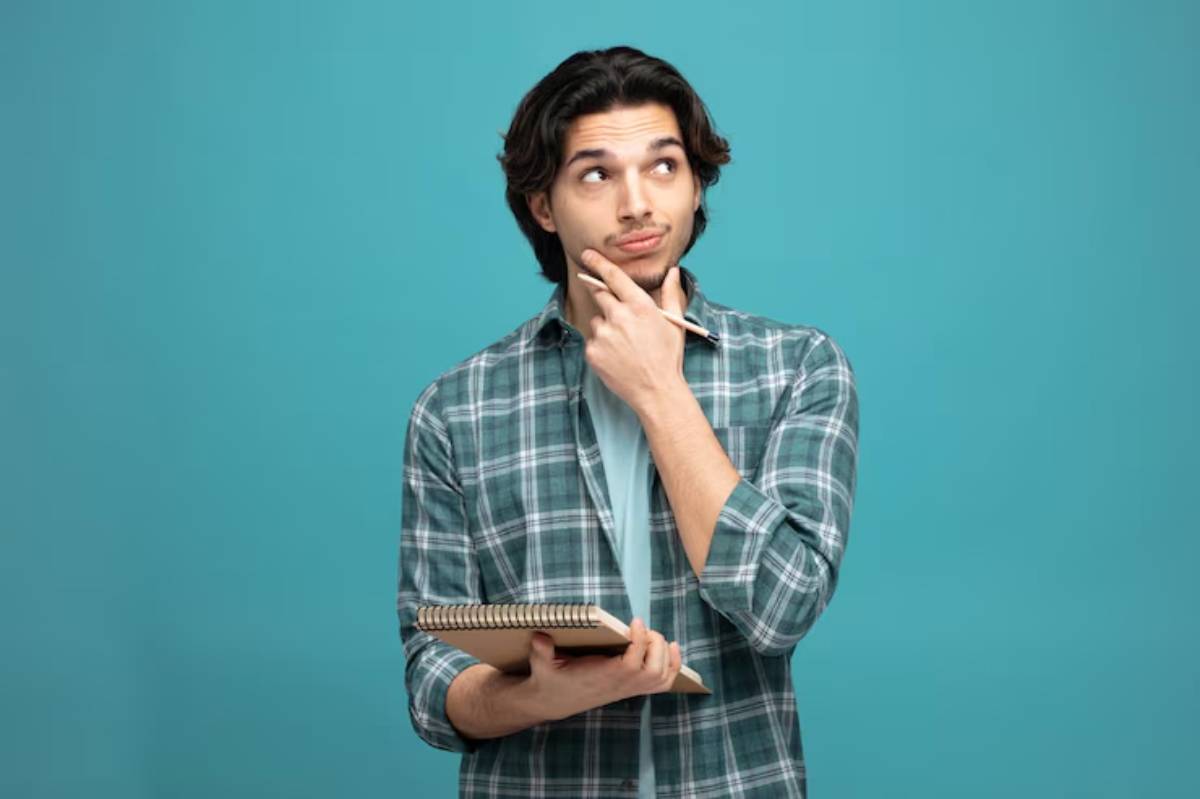 A person in a green plaid shirt stands against a teal background, holding a notebook and a pencil, appearing deep in thought.