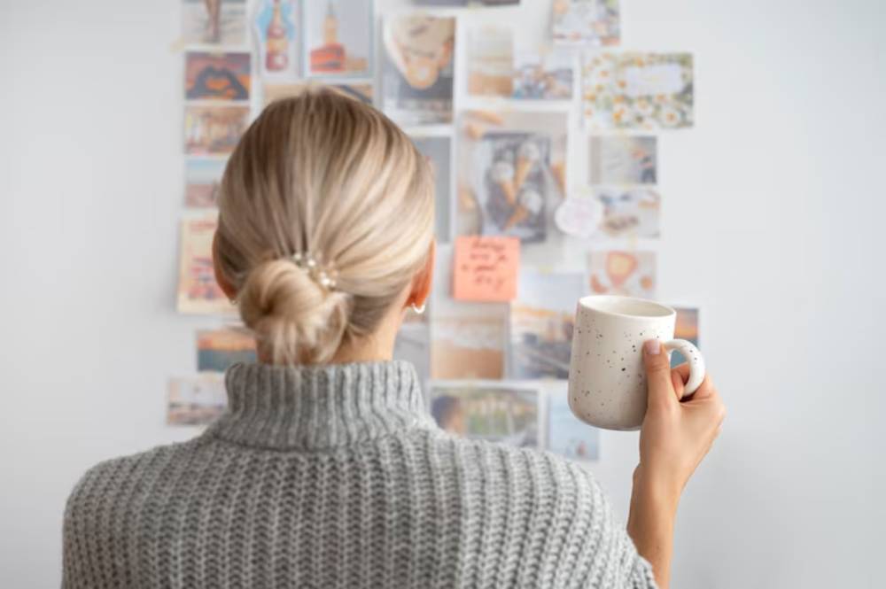 A woman with blonde hair in a bun, wearing a gray sweater, holds a mug and gazes at a vision board on wall.