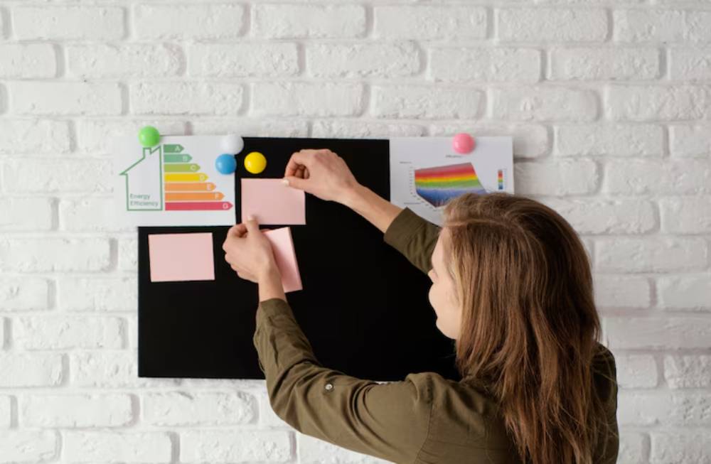 A person arranges pink sticky notes on a black board with energy efficiency charts and colorful magnets attached to a white brick wall.