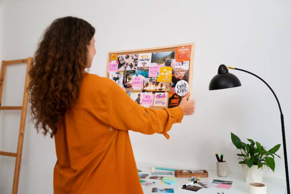 A woman in an orange shirt arranges a vision board filled with inspirational photos and notes near a desk with plants and stationery.