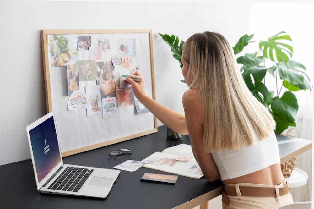 A woman with long hair works at a desk, pinning photos on a corkboard, with a laptop and plants nearby, creating a vision board.