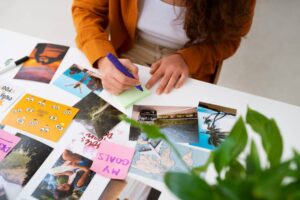 A person writes on a sticky note while arranging photos and goals on a table, surrounded by greenery. Creativity and planning are evident.
